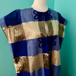 May include: A sleeveless tunic featuring a checkered pattern of metallic gold, royal blue, and light blue squares. The garment has a round neckline with two dark blue decorative frog closures at the center front, creating a vertical slit opening.