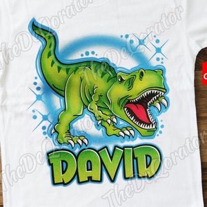 May include: White t-shirt with a green dinosaur graphic and the name "DAVID" in green letters. The dinosaur is roaring with its mouth open and has a red tongue. The dinosaur is surrounded by a blue circle with white stars.