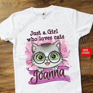 May include: White t-shirt with a pink and purple design featuring a cartoon cat wearing glasses. The text "Just a Girl who loves cats" is printed above the cat. The name "Joanna" is printed below the cat.