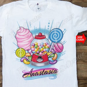 May include: White t-shirt with a colorful candy theme design. The design features a gumball machine, lollipops, jelly beans, and cotton candy. The name "Anastasia" is written in rainbow letters at the bottom of the design.