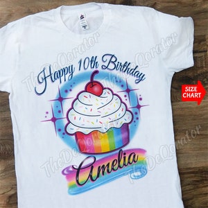 May include: White t-shirt with a colorful airbrushed cupcake design. The cupcake has rainbow frosting, white sprinkles, and a red cherry on top. The text "Happy 10th Birthday Amelia" is written in a cursive font.