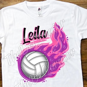 Könnte beinhalten: Weißes T-Shirt mit einem lila und pinkfarbenen Flammen-Design und einem Volleyball. Der Name "Leila" ist in Schwarz über dem Volleyball geschrieben.