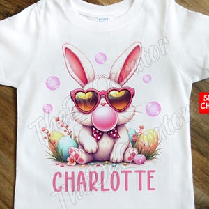 Puede incluir: Camiseta blanca con un conejito de dibujos animados con gafas de sol en forma de corazón y soplando una burbuja. El conejito está rodeado de huevos de Pascua y flores. El nombre "CHARLOTTE" está impreso en rosa.