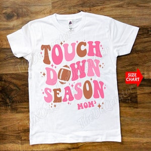 May include: White t-shirt with a pink and brown graphic that says "Touch Down Season" with a football graphic and the text "MOM3".