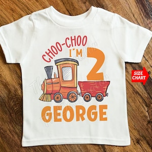 May include: White toddler t-shirt featuring a train graphic. The design includes the text "CHOO-CHOO I'M 2 GEORGE" in orange and red. A red arrow points to a "SIZE CHART" label.