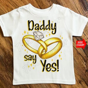 Puede incluir: Camiseta blanca con texto negro que dice "Daddy say Yes!" y un gráfico de dos anillos dorados con un diamante en el centro.