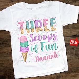 以下が含まれることがあります： ピンク、グリーン、ブルーのアイスクリームコーンのグラフィックが描かれた白いTシャツ。"THREE Scoops of Fun"というテキストがピンク、グリーン、ブルーで書かれています。アイスクリームコーンの下にピンクで"Hannah"という名前が書かれています。