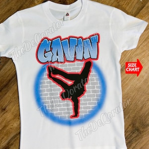 May include: White t-shirt with a blue and gray graffiti design featuring a silhouette of a person breakdancing. The name "GAVIN" is written in a graffiti style above the design.