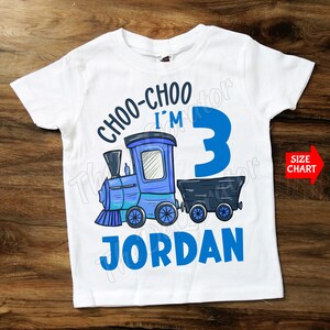 May include: White t-shirt featuring a blue train graphic and the text "CHOO-CHOO I'M 3 JORDAN". The train is blue with black accents. A red arrow with "SIZE CHART" is on the right side.