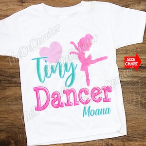 May include: White t-shirt with a pink heart, a teal blue "tiny dancer" text, and a pink silhouette of a ballerina. The name "Moana" is written in teal blue below the text.