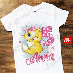 Può includere: Maglietta bianca con un gatto cartone animato rosa e giallo che indossa un fiocco rosa, il numero 5 con pois rosa e il nome "Anna" in lettere rosa.