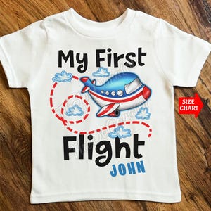 May include: White t-shirt with a colorful airplane graphic. The design includes the text "My First Flight" in black, with the name "John" in blue. A red arrow points to a "Size Chart" label.
