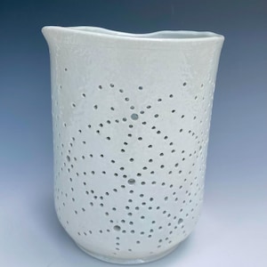 Könnte beinhalten: Eine weiße Keramikvase mit strukturierter Oberfläche und einer Reihe kleiner, gleichmäßig verteilter Löcher. Die Vase hat einen leicht unregelmäßigen Rand und eine zylindrische Form, ideal für Blumen oder als dekoratives Element. Höhe: ca. 20 cm.
