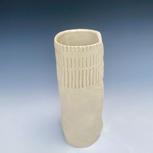 May include: A tall, off-white ceramic vase with a rectangular opening. The upper portion features vertical, textured lines, while the lower section has a swirling pattern. The vase is approximately 8 inches tall.