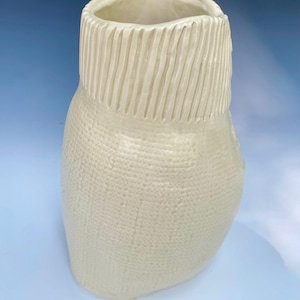 May include: Off-white ceramic vase with a textured surface. The top section features vertical ribbed detailing, while the body has a dotted pattern. The vase has a wide opening and a rounded base. This decorative vase is suitable for home decor.
