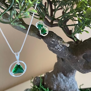 May include: A silver necklace with a green triangular pendant set in a circle. The pendant is surrounded by a silver circle. The necklace is hanging from a small green triangular earring.