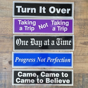 May include: Five rectangular bumper stickers with white text on coloured backgrounds. The stickers read: Turn It Over, Taking a Trip Not Taking a Trip, One Day at a Time, Progress Not Perfection, and Came, Came to Came to Believe.