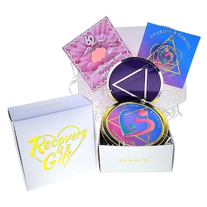 May include: A white box with the text "Recovery is a Gift" in gold lettering. Inside the box are several medallions and cards with the words "Sharing & Caring" and "Sponsor". The medallions have a heart and triangle design.