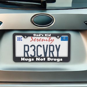 May include: A license plate with a black frame is mounted on a car. The plate reads "R3CVRY" in bold blue letters, with "Hugs Not Drugs" below. Above the plate, the frame reads "God's Kid" and "Serenity".