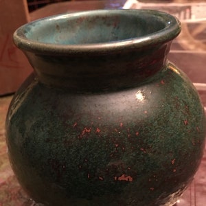 May include: A green ceramic vase with a speckled glaze and a copper-colored hue. The vase has a round body and a wide opening.