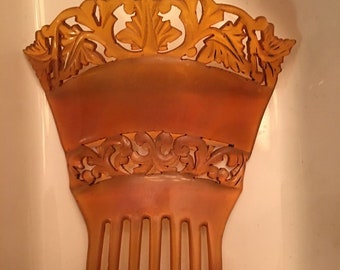May include: A vintage amber-colored plastic hair comb with a decorative floral design. The comb has a wide, curved top with intricate cutouts and a row of seven teeth.