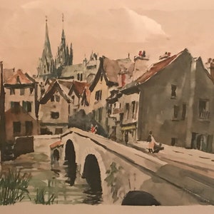 May include: A watercolor painting of a European town with a bridge over a river. The buildings are in shades of brown and gray, and the sky is a light blue. There are trees in the background, and a church with two tall spires is visible in the distance.