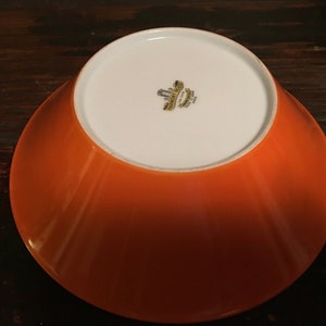 May include: Orange ceramic bowl with a white interior. The bowl has a raised rim and a smooth, glossy finish. The bottom of the bowl has a gold sticker with the text "Made in Japan".