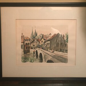 May include: A framed watercolor painting depicting a European village scene with a bridge over a river. The buildings are painted in shades of brown, gray, and white, with a church steeple in the background. The painting is set against a light blue sky.