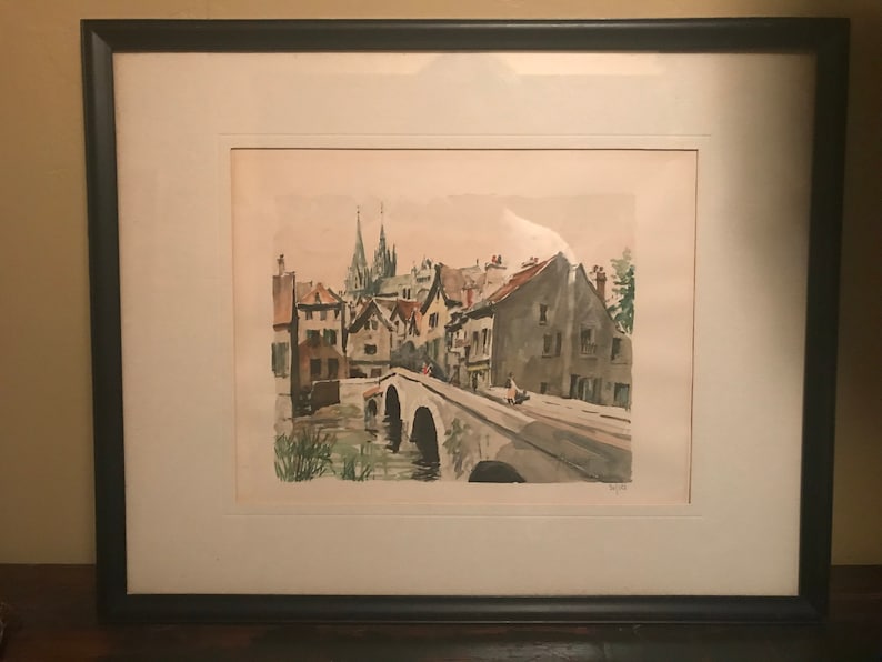May include: A framed watercolor painting depicting a European village scene with a bridge over a river. The buildings are painted in shades of brown, gray, and white, with a church steeple in the background. The painting is set against a light blue sky.