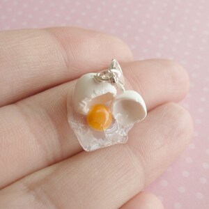 May include: A white and yellow charm in the shape of a cracked egg with a yolk inside. The charm is made of polymer clay and is attached to a silver chain.