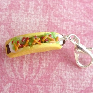 May include: A miniature hot dog charm with a yellow bun, white sauce, green lettuce, red ketchup, and yellow cheese. The charm has a silver lobster clasp.