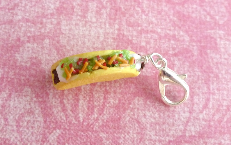 May include: A miniature hot dog charm with a yellow bun, white sauce, green lettuce, red ketchup, and yellow cheese. The charm has a silver lobster clasp.