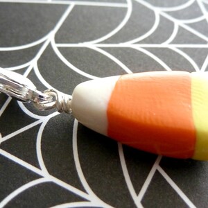May include: A silver charm in the shape of a candy corn with white, orange, and yellow stripes. The charm is attached to a silver loop.