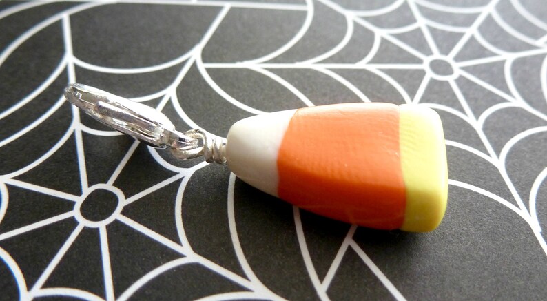 May include: A silver charm in the shape of a candy corn with white, orange, and yellow stripes. The charm is attached to a silver loop.