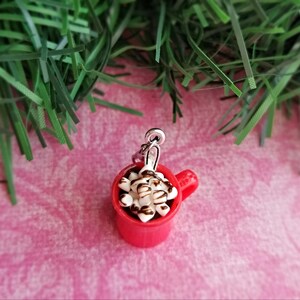May include: A miniature red mug charm filled with white marshmallows and drizzled with chocolate. The charm has a silver-colored loop for attaching to a bracelet or necklace. The background is a pink surface with green decorative accents.