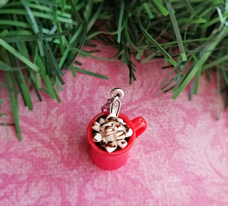May include: A miniature red mug charm filled with white marshmallows and drizzled with chocolate. The charm has a silver-colored loop for attaching to a bracelet or necklace. The background is a pink surface with green decorative accents.