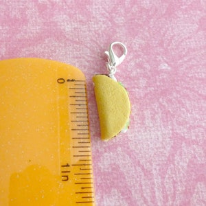 May include: A small, yellow taco charm with a silver clasp. The taco is filled with lettuce, tomato, and cheese.
