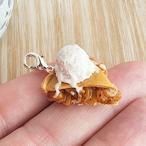 May include: A silver charm in the shape of a slice of pie with a dollop of white frosting and a scoop of white ice cream on top.