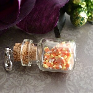May include: A small, clear glass bottle filled with candy corn. The bottle has a cork stopper and a silver-colored clasp. The candy corn is orange, yellow, and white. The background includes a purple flower and a patterned surface.