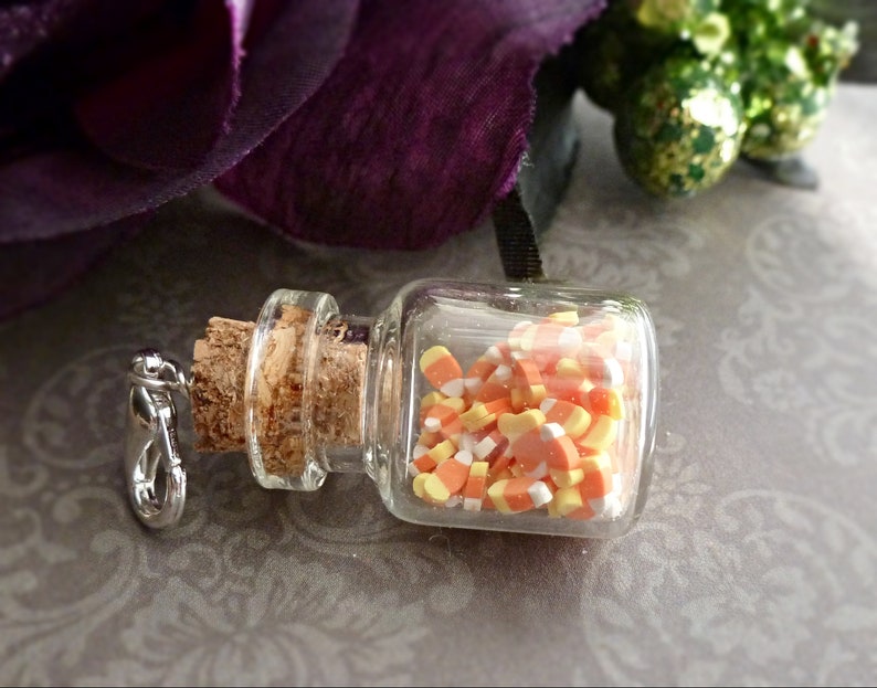 May include: A small, clear glass bottle filled with candy corn. The bottle has a cork stopper and a silver-colored clasp. The candy corn is orange, yellow, and white. The background includes a purple flower and a patterned surface.