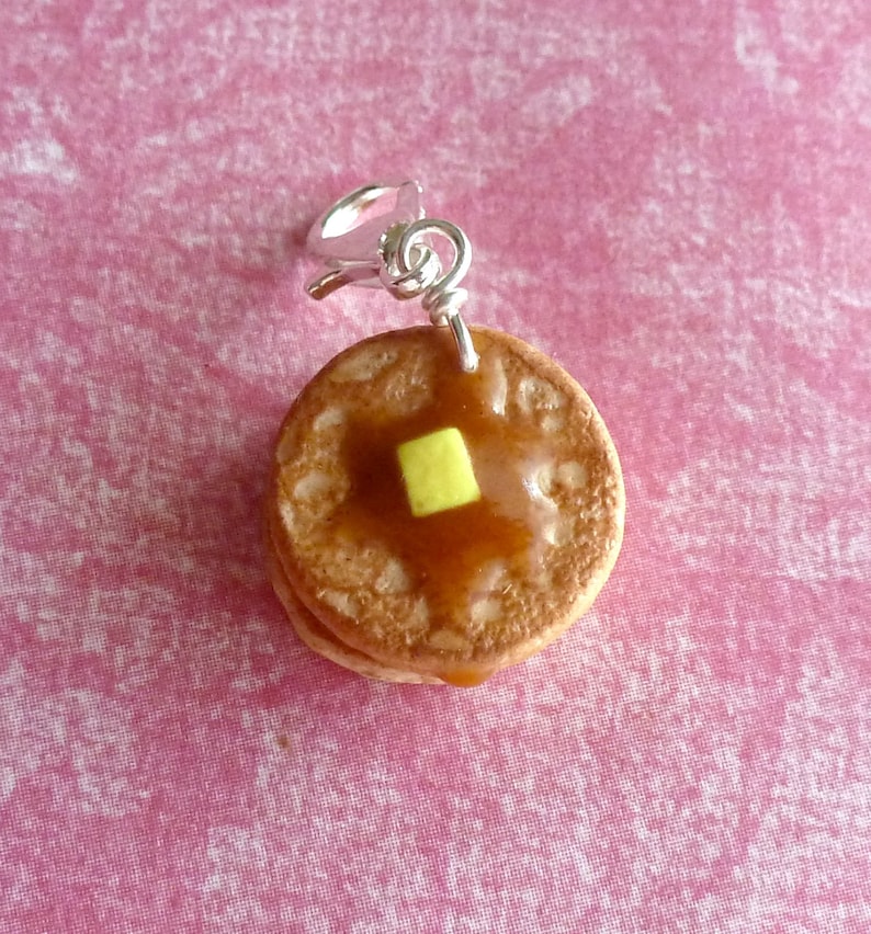 May include: A small, brown, pancake charm with a yellow pat of butter and brown syrup on top. The charm is attached to a silver jump ring.
