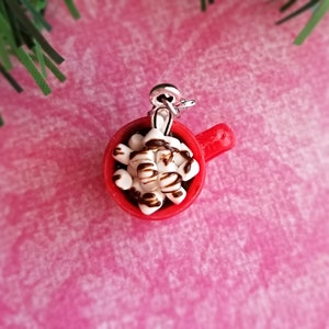 May include: A miniature red mug charm filled with white marshmallows drizzled with chocolate. The charm has a silver-colored loop for attaching to a necklace or bracelet. The background is a pink textured surface with a sprig of green.