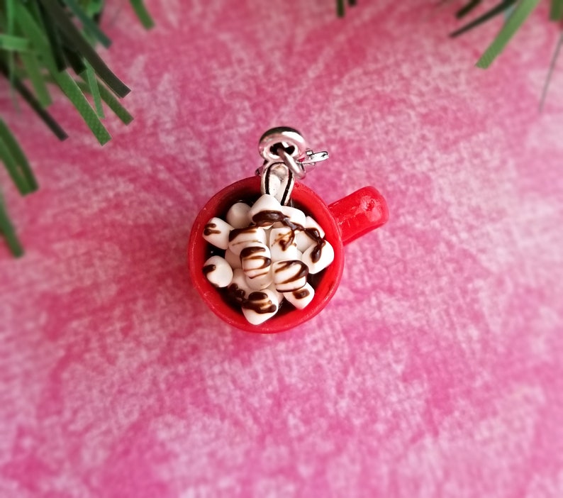 May include: A miniature red mug charm filled with white marshmallows drizzled with chocolate. The charm has a silver-colored loop for attaching to a necklace or bracelet. The background is a pink textured surface with a sprig of green.