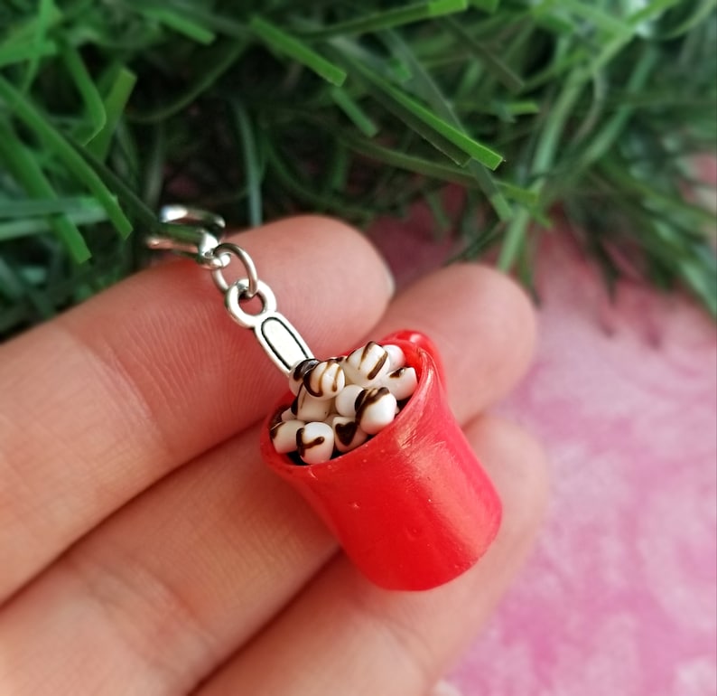 May include: A miniature red mug filled with white marshmallows drizzled with chocolate. The mug has a silver handle and is attached to a silver clasp. The charm is a festive accessory.