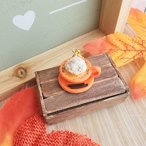 May include: A miniature orange ceramic mug with whipped cream and a sprinkle of cinnamon, sitting on a wooden crate. The mug is a charm or keychain.