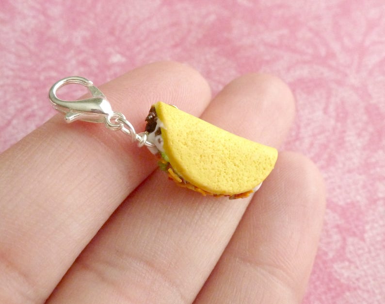 May include: A silver charm in the shape of a taco. The taco is filled with meat, cheese, and lettuce. The charm is attached to a silver clasp.