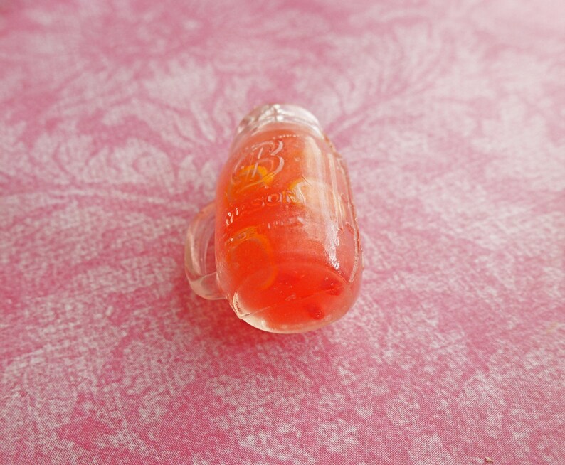 May include: A clear glass jar with a red liquid and a yellow citrus fruit inside. The jar has a handle and a label that reads "Ball."