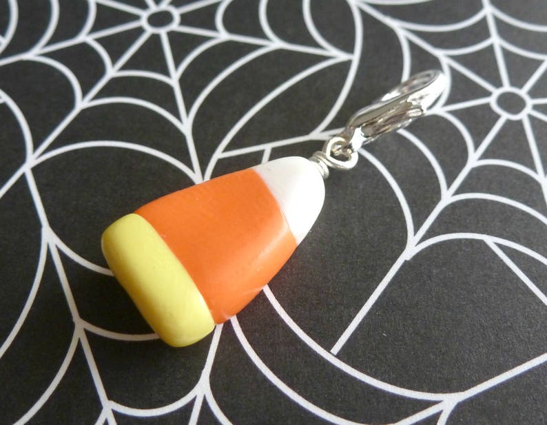 May include: A silver charm in the shape of a candy corn with orange, yellow, and white stripes. The charm is attached to a silver chain.