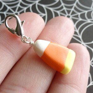 May include: A silver charm in the shape of a candy corn with white, orange, and yellow stripes. The charm is attached to a silver jump ring.
