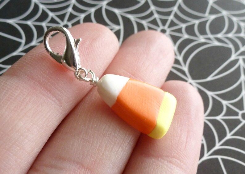 May include: A silver charm in the shape of a candy corn with white, orange, and yellow stripes. The charm is attached to a silver jump ring.